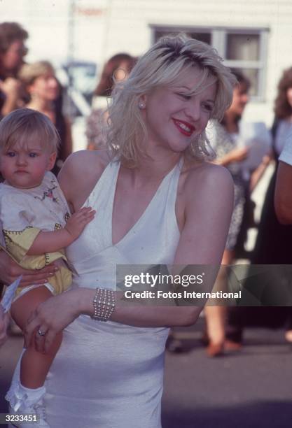 American rock singer Courtney Love, wearing a white satin dress, smiles while holding her daughter by Kurt Cobain, Frances Bean, outside the 1993 MTV...