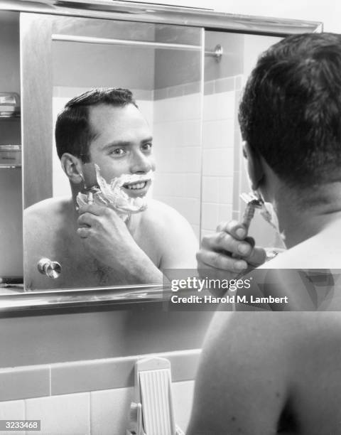 Shirtless man with shaving cream on his face looks into a bathroom mirror as he shaves with a safety razor.