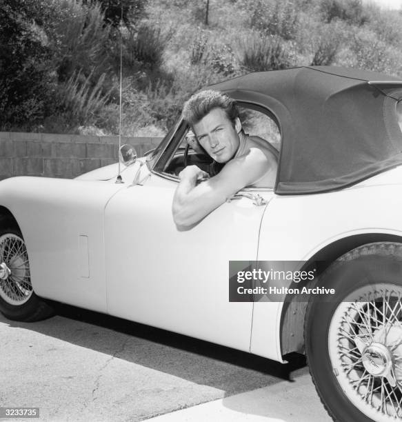 Portrait of American actor Clint Eastwood leaning out the window of a Jaguar convertible without a shirt.