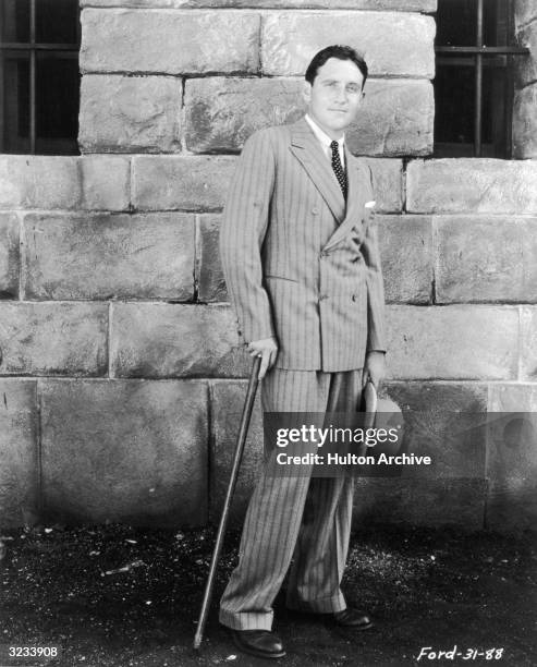 Full-length, outdoor portrait of American actor Spencer Tracy wearing a pinstriped, double-breasted suit, holding a cane and a hat.