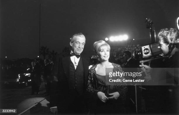 American actor and film director John Huston smiles with American actor Stella Stevens as a television cameraman films them arriving at the Academy...
