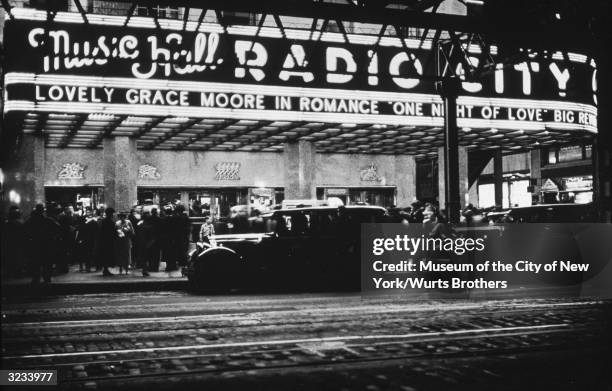 Cars stand in front of a crowded Radio City Music Hall, with a marquee advertising Gracie Moore in the film, 'One Night of Love,' New York City.