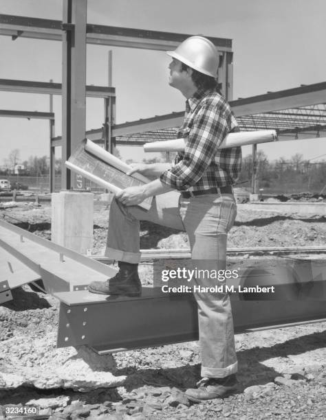 Construction worker at a construction site rests a boot on an I-beam while reviewing blueprints.
