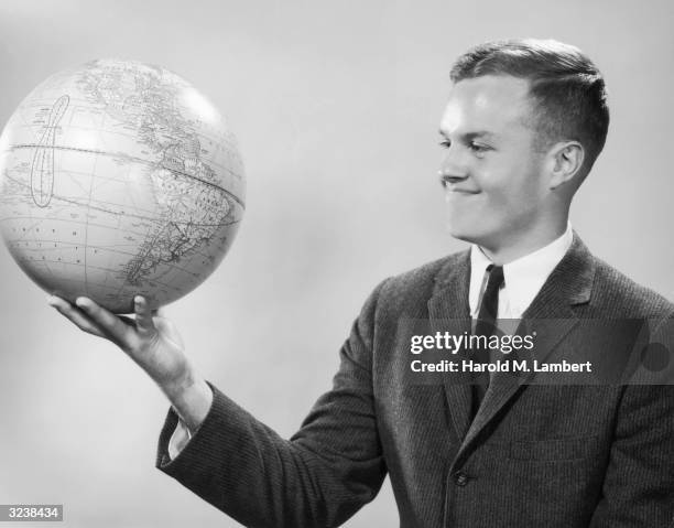 Studio portrait of a young man, wearing a suit and tie, holding a globe in one hand.
