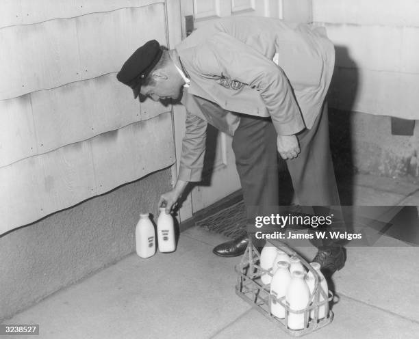 Milkman wearing his delivery uniform bends down as he leaves two bottles of milk by the front door of a house. There is a metal basket full of milk...