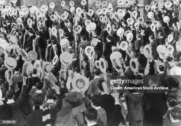 Overhead view of a crowd of young men standing outdoors, waving their straw hats in the air.
