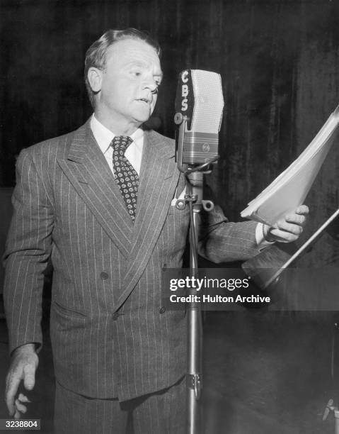 American actor James Cagney , wearing a double-breasted pinstripe suit, stands and reads from a script into a CBS radio microphone at a studio.