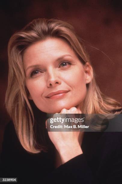 Headshot of American actor Michelle Pfeiffer smiling while resting her chin on her hand.