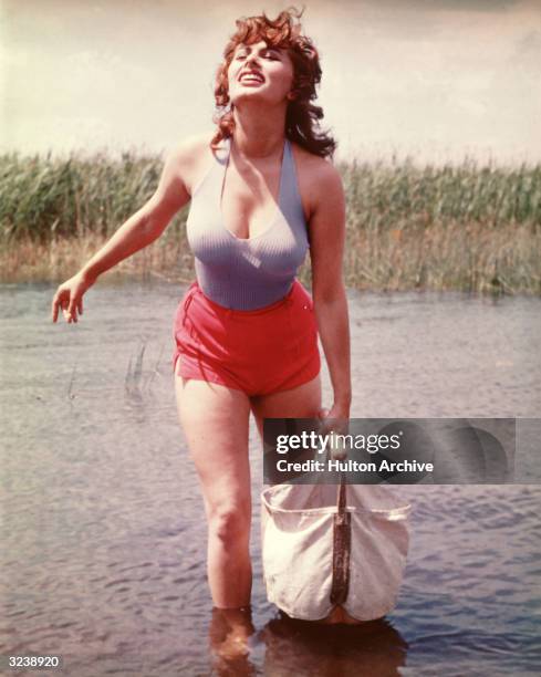 Italian actor Sophia Loren, wearing low-cut lavender tank top and red hot shorts, carries a canvas bag while crossing a stream.