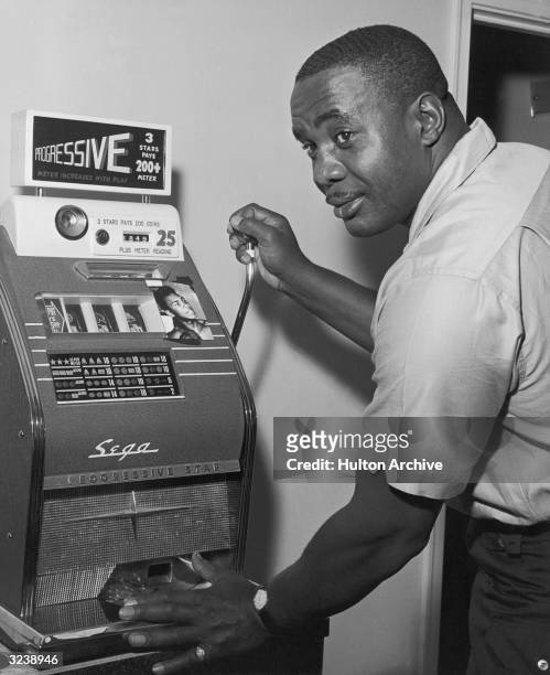 American heavyweight champion boxer Sonny Liston looks over his shoulder while pulling the handle of a slot machine to which a small photograph of...
