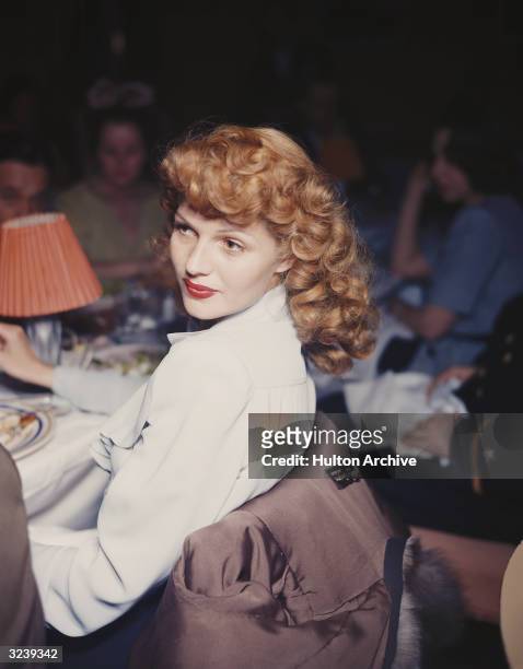 American actor Rita Hayworth turns her head while leaning back in a chair at a dining table in a restaurant. Hayworth wears a white blouse with...