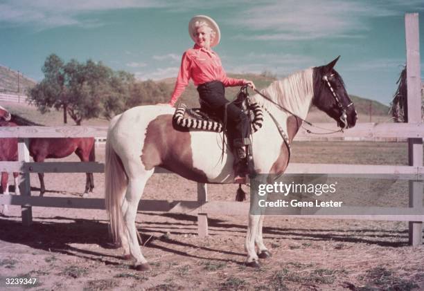 Full-length portrait of American actor Betty Grable squinting in the sun while seated on a pinto horse with brown patches in a corral. Grable wears a...