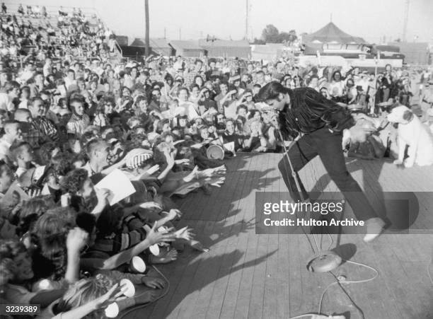 American singer and actor Elvis Presley performing outdoors on a small stage to the adulation of a young crowd.