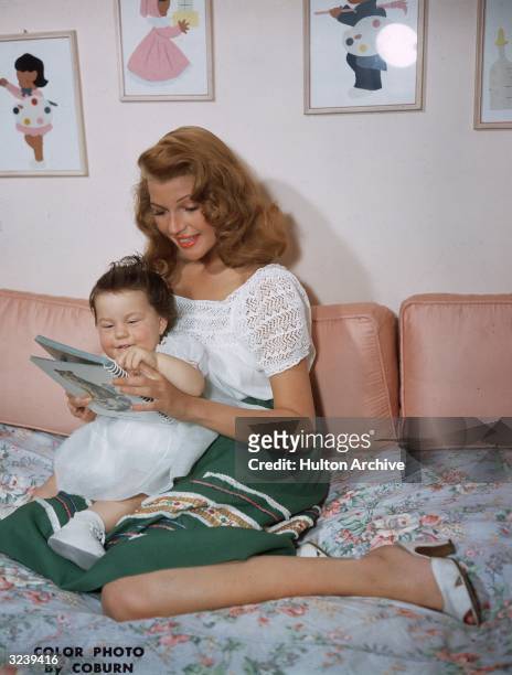 American actor Rita Hayworth looks through an illustrated book with her infant daughter, Rebecca Welles, as she holds her in her lap while sitting on...