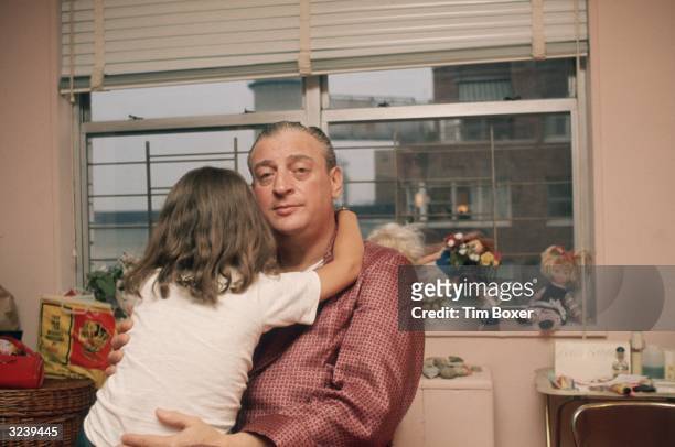 American comedian and actor Rodney Dangerfield, dressed in silk pajamas, holds his daughter as she hugs his neck and turns her back to the camera,...