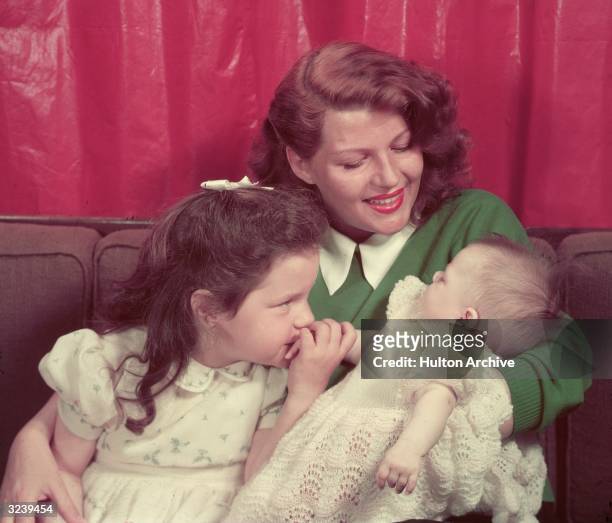 Portrait of American actor Rita Hayworth sitting on a sofa with her young daughters, Rebecca Welles and Yasmin Khan, in her lap. Hayworth is wearing...