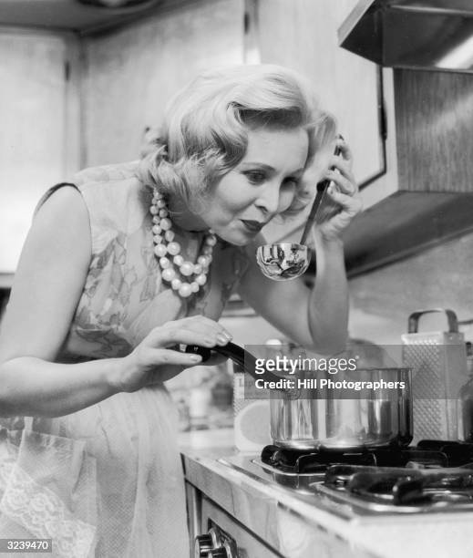 Housewife tasting the soup she is cooking for dinner by dipping a ladle into the saucepan on the hob.