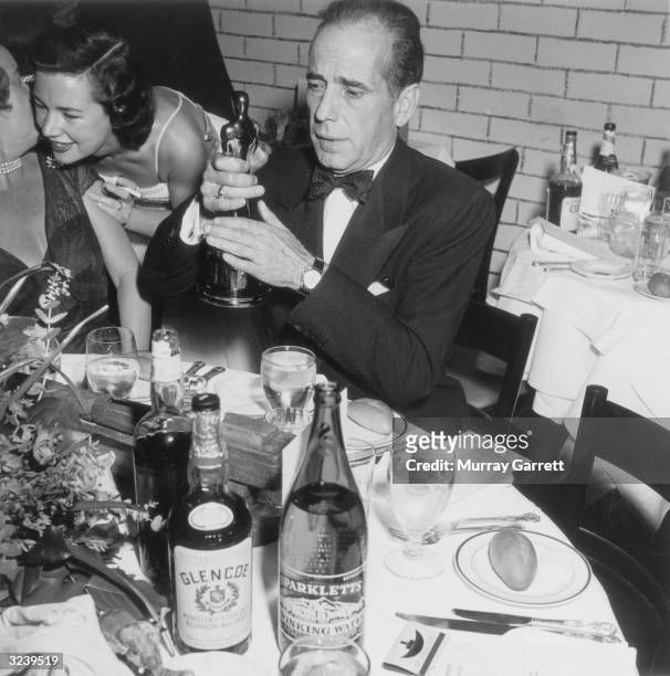 American actor Humphrey Bogart sits at a table with place settings and liquor bottles, holding up his Best Actor statue at the Academy Awards, RKO...