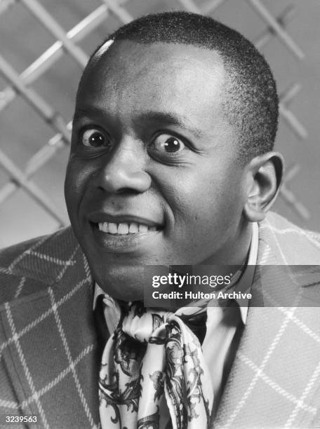 Headshot portrait of American comedian and actor Flip Wilson , wearing a blazer with a silk scarf tied around his neck, making a face for the camera.