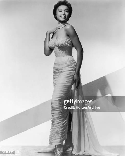 Full-length studio portrait of American actor Diahann Carroll wearing an evening gown with a long sash. She holds one hand by her shoulder.