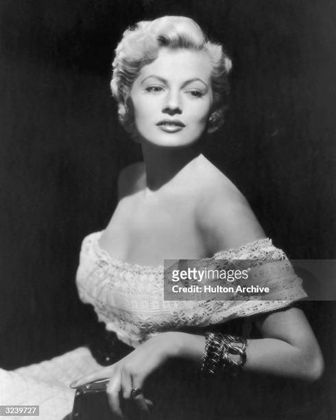 Promotional portrait of Swedish-born actor Anita Ekberg sitting on a chair and wearing an off-the-shoulder crocheted dress, with a black backdrop.