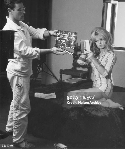 French actor Brigitte Bardot uses a mirror while a crew member holds the clapperboard in front of her on the set of director Jean-Luc Godard's film,...