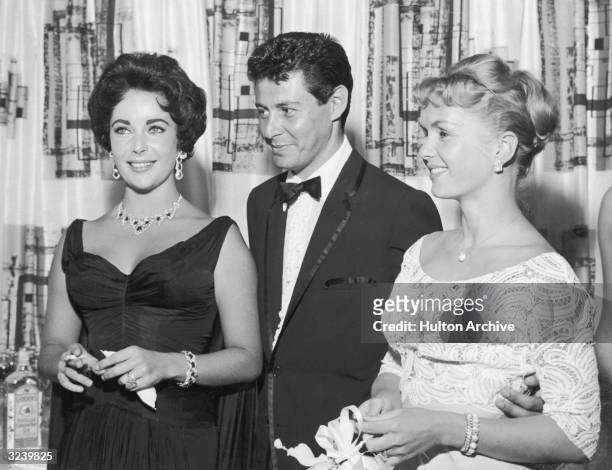 American singer Eddie Fisher, wearing a tuxedo, stands with arm around his wife, American actor Debbie Reynolds and smiles while looking at...