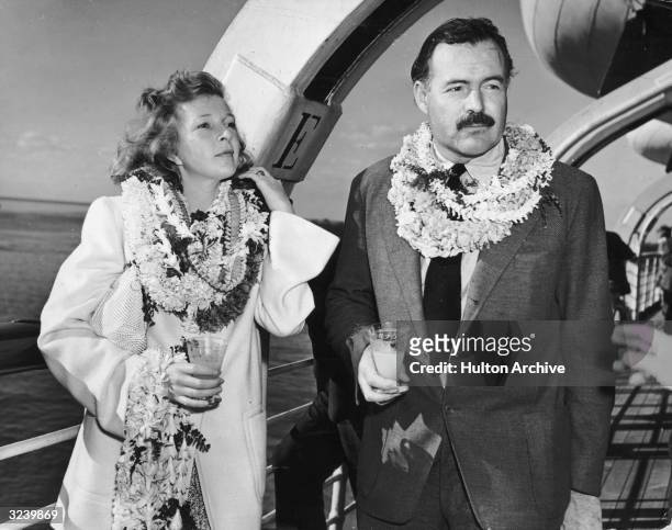 American author Ernest Hemingway and his wife, journalist Martha Gellhorn , stand on deck aboard a ship, wearing multiple leis and holding cocktails.