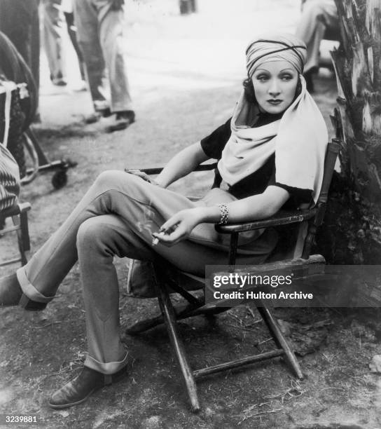 German-born actor Marlene Dietrich sits in a canvas chair wearing a turban and holding a cigarette on the desert location set of director Richard...