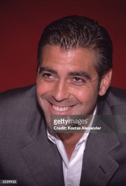 Studio headshot of actor George Clooney smiling and leaning forward in front of a red backdrop.