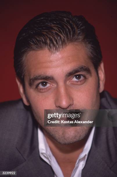 Headshot of American actor George Clooney with stubble, in front of a red background.