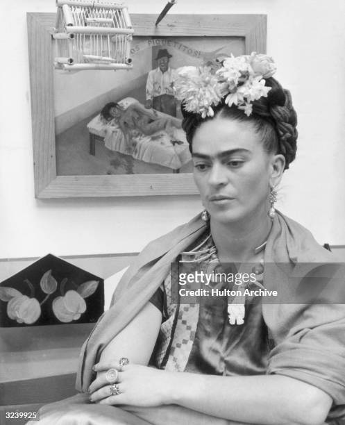 Mexican painter Frida Kahlo sits with her arms folded, looking down, in front of one of her paintings and a wooden bird cage. She wears flowers in...