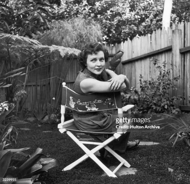 American actor Betty White sits in a canvas chair with her name written on the back, looking over her shoulder in a backyard garden.