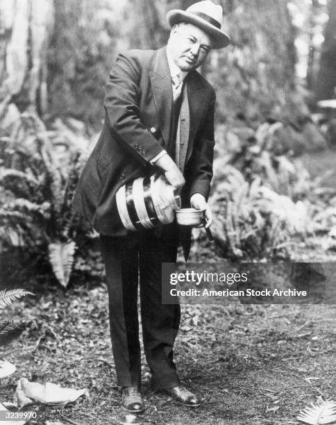 American president Herbert Hoover pours a jug and smokes a cigar in a wooded area.