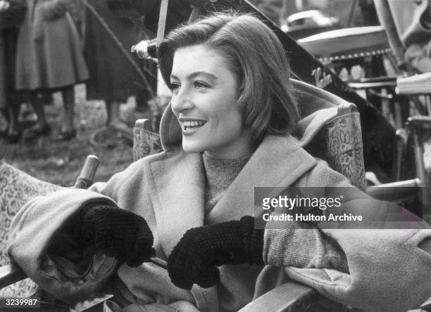 French actor Anouk Aimee, wearing a camel-hair overcoat and gloves, laughs while sitting in a folding chair on a film set.