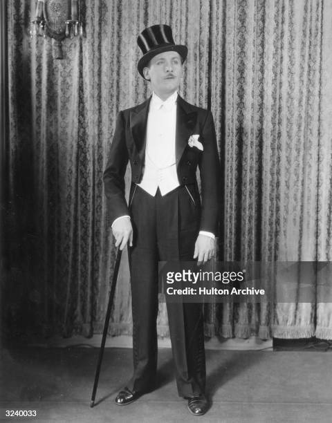 Full-length portrait of a man standing in front of a curtain wearing coattails, white tie, and a top hat while holding a cane.