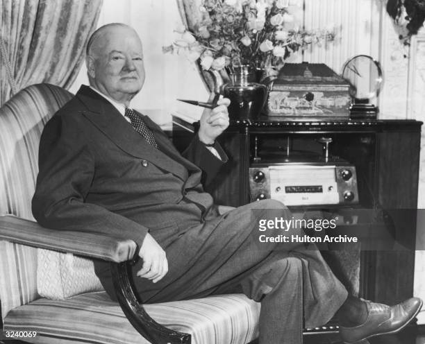 Portrait of former American president Herbert Hoover seated in an armchair with a pipe in his suite at the Waldorf Towers, New York City.