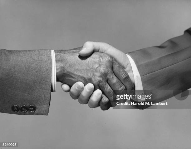 Close-up of a dark skinned hand clasping a light skinned hand in a handshake, 1970s.