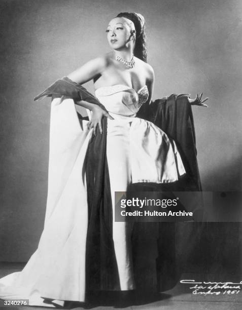 Full-length studio portrait of American singer and performer Josephine Baker , wearing a fall with her hair pulled back, standing in a strapless gown...