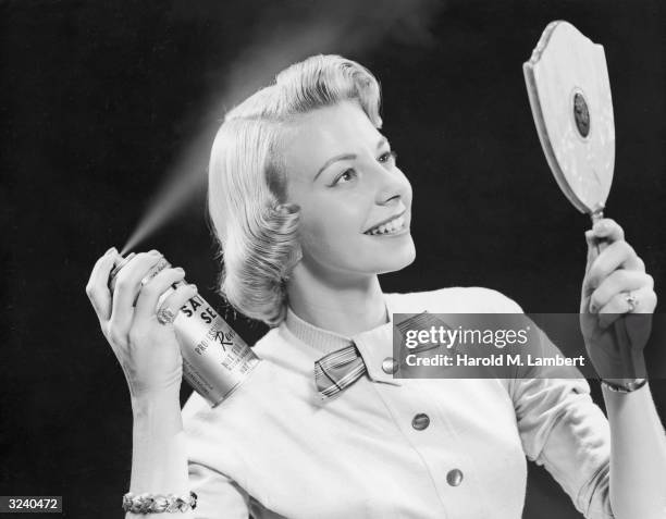 Young woman uses a hand-held mirror to apply Revlon hair spray to her hair.