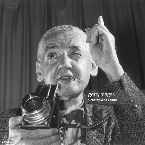 Headshot of Polish-born photojournalist Arthur Fellig , known as 'Weegee', holding a camera and pointing his index finger, with a cigar in his mouth.