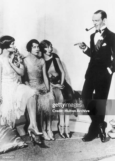 American actor, singer, and dancer Fred Astaire leans against a wall with a pipe in his mouth, pointing as American actors : Betty Compton, Adele...