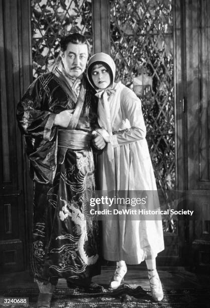 Swedish actor Warner Oland, made up to appear Asian and wearing a kimono, stands in front of a door, arm in arm with Russian actor Dorothy Green, in...