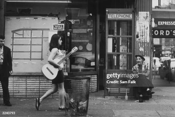 Street scene of a young woman walking with an acoustic guitar, as an old man sits by a telephone booth, in Greenwich Village, New York City. A man...