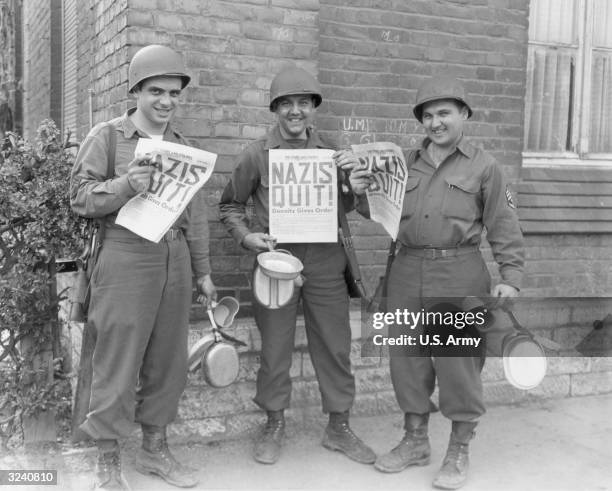 Full-length portrait of three United States Ninth Army soldiers smiling as they hold up copies of 'The Stars and Stripes' announcing Germany's...