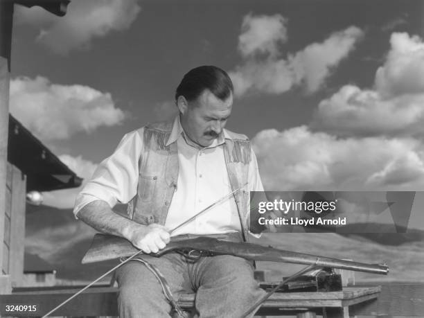 American writer Ernest Hemingway sits outdoors and cleans his Mannlicher rifle, a gun he first used during his African hunts, Idaho.