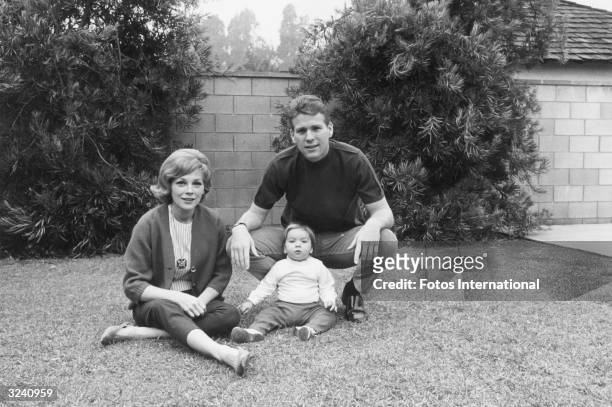 American actor Ryan O'Neal with his wife, actor Joanna Moore, and daughter, Tatum, sitting on grass in front of a stone wall.