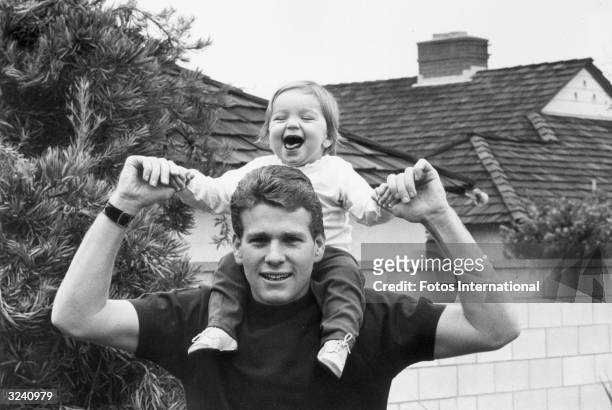 American actor Ryan O'Neal holding his smiling daughter Tatum on his shoulders in front of a house.