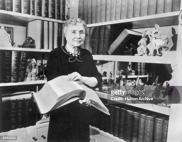 Portrait of American writer, educator and advocate for the disabled Helen Keller holding a Braille volume and surrounded by shelves containing books...
