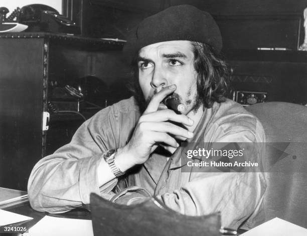 Argentine-born Cuban revolutionary leader and economic advisor Ernesto 'Che' Guevara sits at a desk and smokes a cigar, wearing military fatigues and...